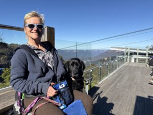 Woman smiling at camera next to black labrador with blue sky behind them